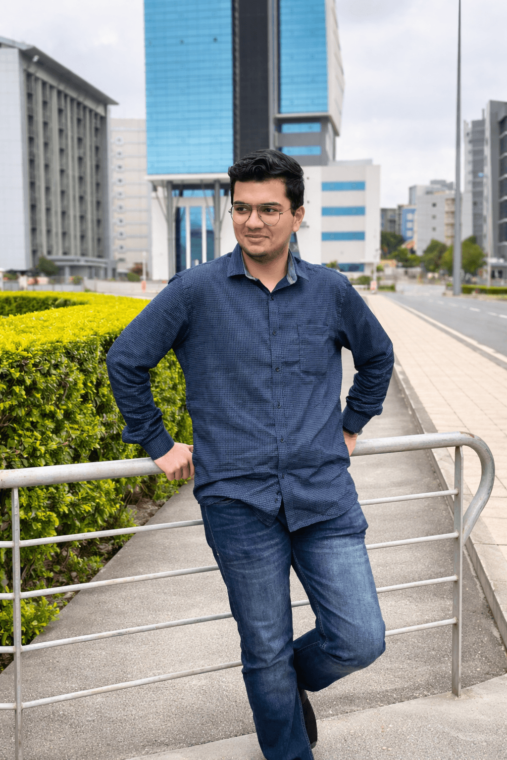 Man in glasses and blue shirt leaning on a railing with modern city buildings behind.